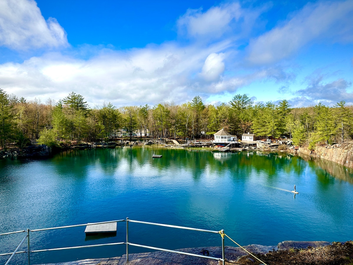 A serene view of a crystal-clear spring-fed quarry is captured, surrounded by granite cliffs and lush trees. In the distance, the Cabana is nestled among the greenery, with a deck extending over the water. The peaceful surface reflects the blue sky and clouds.