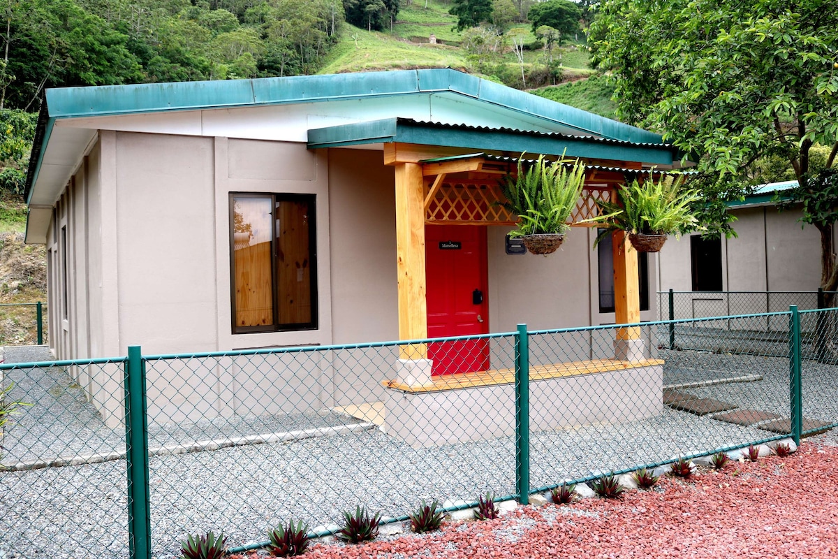 The exterior of a single-story villa is shown, featuring a red door framed by wooden accents. Flanking the entrance are hanging planters. A gravel pathway leads to the entrance, surrounded by a decorative fence. Lush green hills are visible in the background.