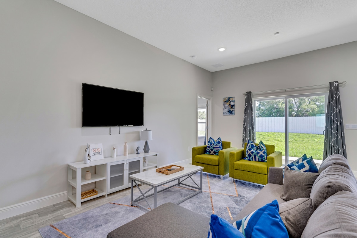A spacious living room features a large grey sectional sofa and two vibrant green chairs. A flat-screen TV is mounted on the wall above a low coffee table. Natural light filters through sliding glass doors, revealing a serene outdoor view.