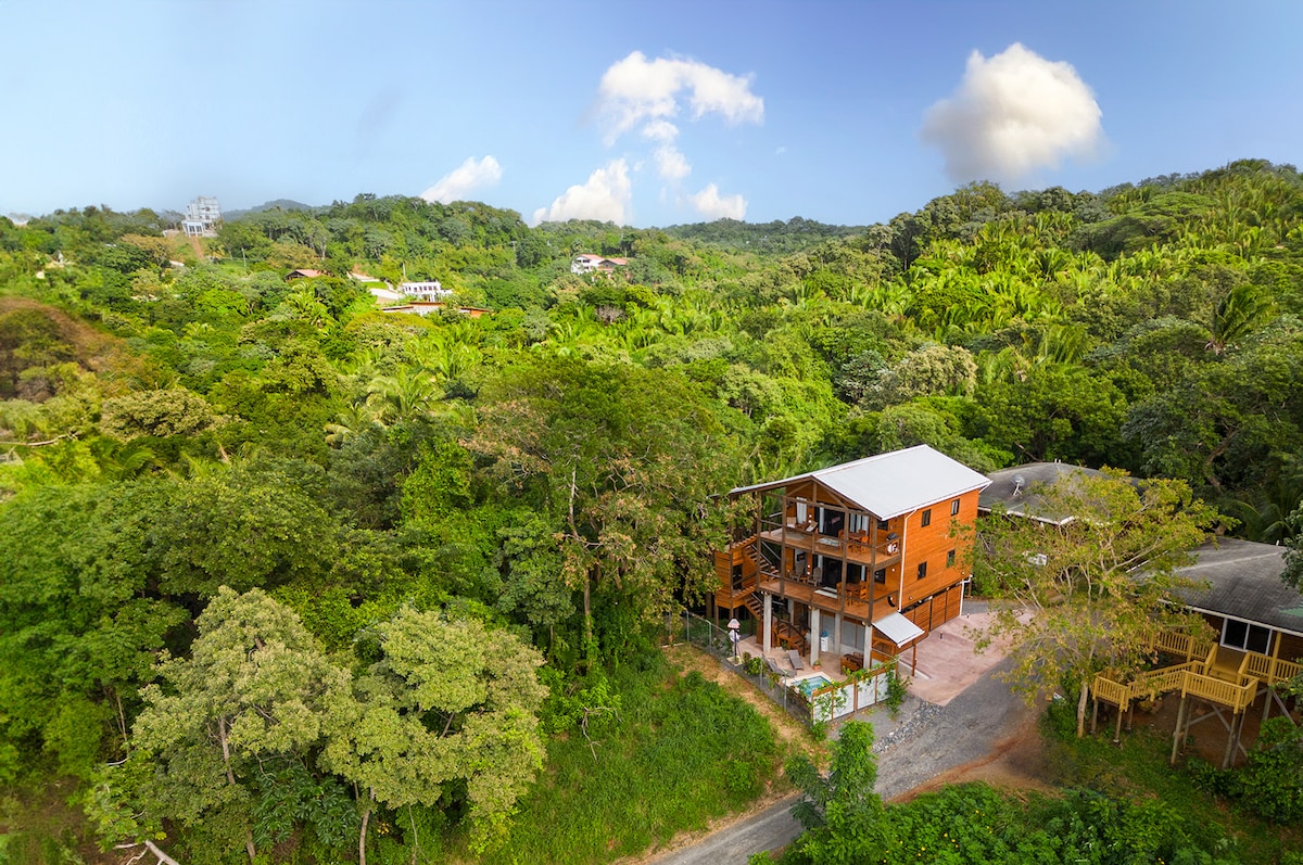 The building is set in a lush, green landscape, surrounded by dense trees and tropical vegetation. The structure's three levels are visible, demonstrating a blend of wood and modern design. Nearby, rolling hills and a clear blue sky complement the scenic surroundings.