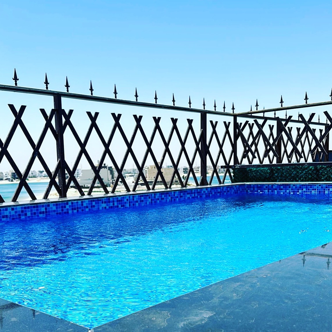 A refreshing swimming pool is visible, featuring clear blue water under a bright sky. The area is enclosed by a decorative railing with a crisscross pattern, providing a sense of privacy while allowing for scenic views of the surrounding landscape.
