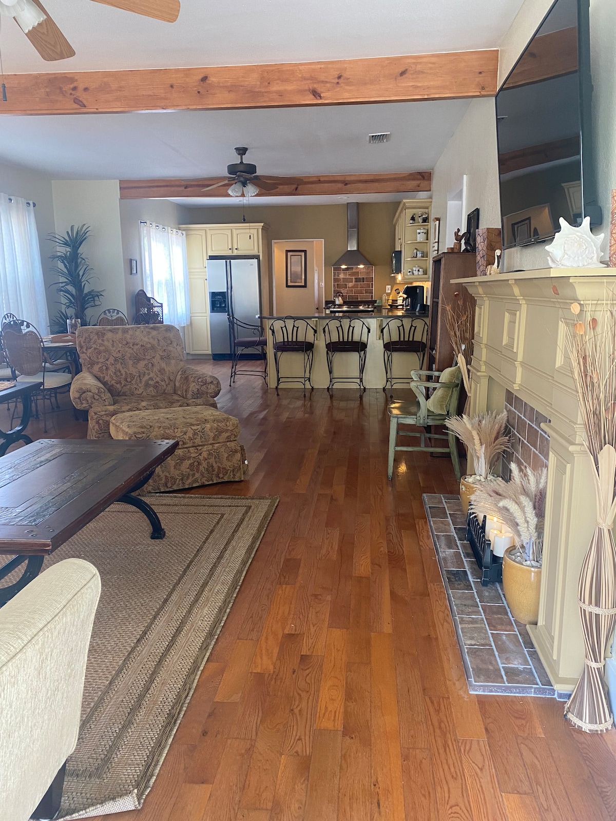 A spacious living area features warm wooden floors and exposed beams. A comfortable armchair is positioned near a fireplace, while a dining area with a large table is visible in the background. A well-equipped kitchen can be seen beyond the dining area.
