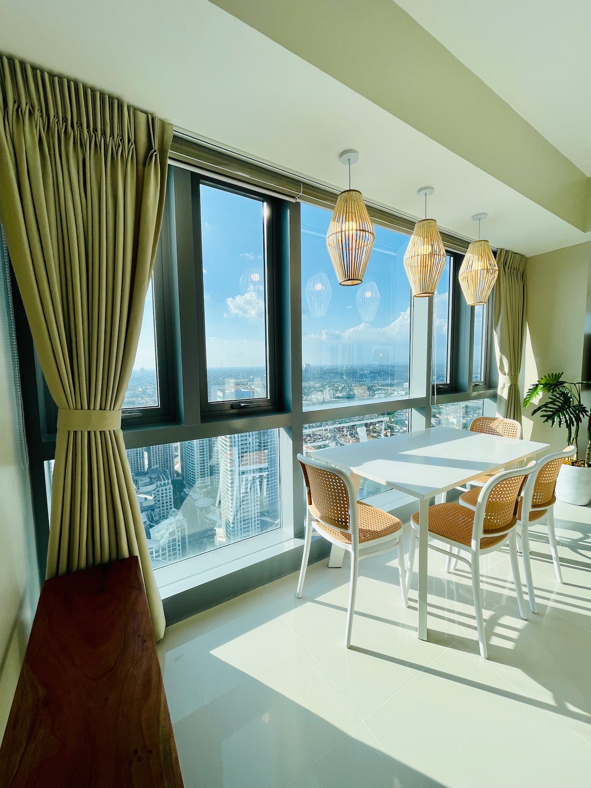A dining area is illuminated by natural light from large floor-to-ceiling windows, offering breathtaking city views. A modern white table is surrounded by four chairs, with decorative pendant lights hanging above. Soft beige curtains complement the light, airy ambiance.