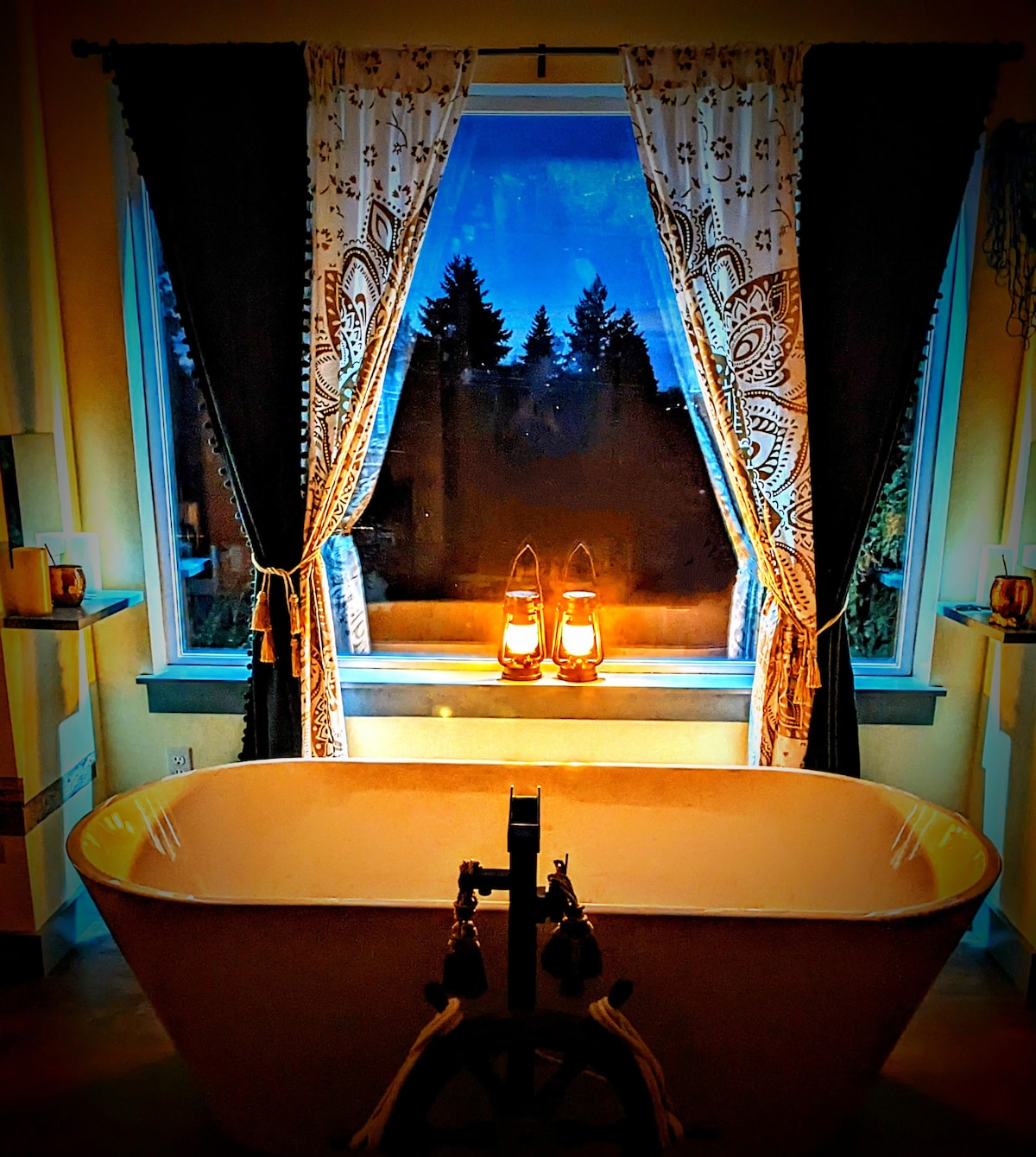 A luxurious soaking tub is positioned in front of a large window, framed by decorative curtains. Two lanterns provide soft illumination. Outside the window, trees are silhouetted against a twilight sky, enhancing the serene ambiance of the bathroom.