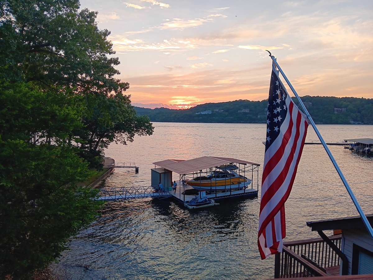 A serene lakeside scene is captured at sunset, with gentle waves reflecting hues of orange and pink in the water. A covered boat slip is visible, housing a boat, while an American flag gracefully waves nearby against the backdrop of lush greenery and a distant shoreline.