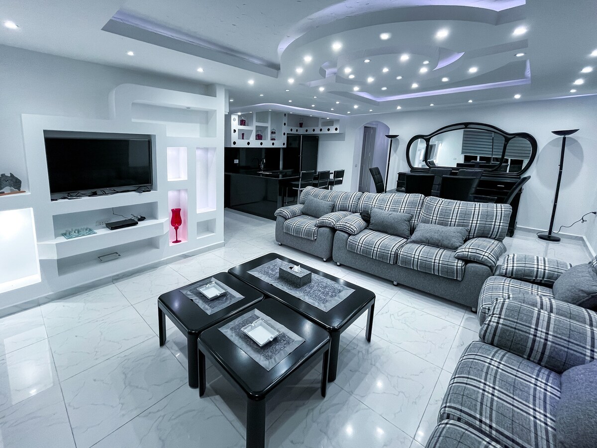 A modern living area features a comfortable gray plaid sectional sofa paired with a sleek black coffee table. A wall-mounted television and soft ambient lighting complement the space, while an open dining area with a dark wood table and chairs is visible in the background.