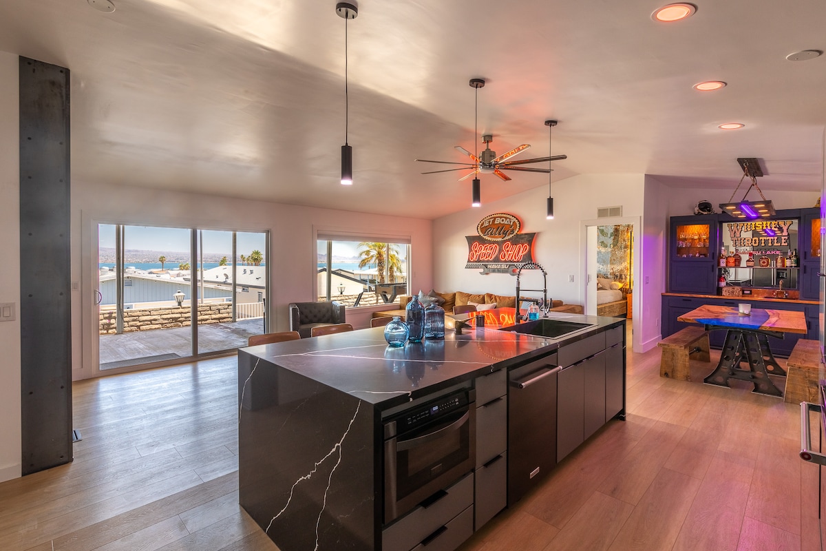 A modern kitchen is visible, featuring a sleek island with black stone countertops and stainless steel appliances. The open space leads to large windows showcasing views of Lake Havasu. A cozy seating area is set behind, illuminated by natural light.