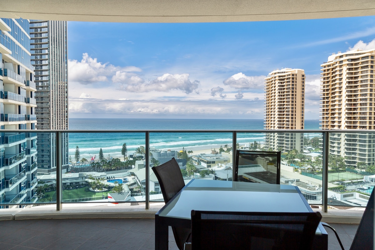 A spacious balcony offers stunning views of the blue ocean and beach. A glass table with black chairs is positioned for outdoor seating. In the background, the towering buildings of Surfers Paradise create a striking contrast against the sky.
