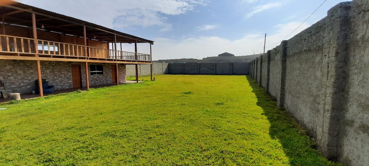 A spacious outdoor area is presented, featuring well-maintained green grass under a clear sky. A two-story structure with wooden balconies is visible in the background, accompanied by a solid wall that encloses the space, ensuring privacy.