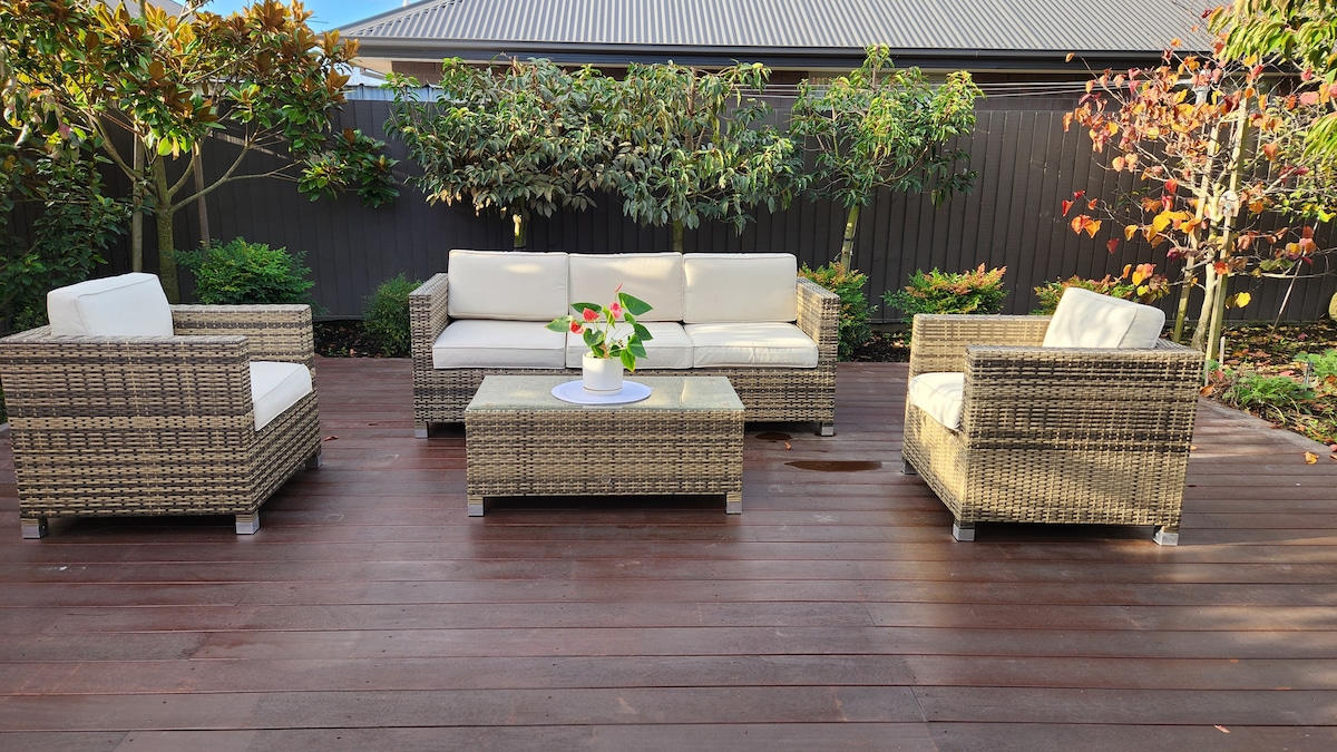 An outdoor seating area features a set of woven brown rattan furniture, including a spacious sofa and two armchairs, arranged around a glass coffee table. Surrounding greenery and seasonal foliage enhance the relaxed atmosphere, while wooden decking provides a sturdy foundation.