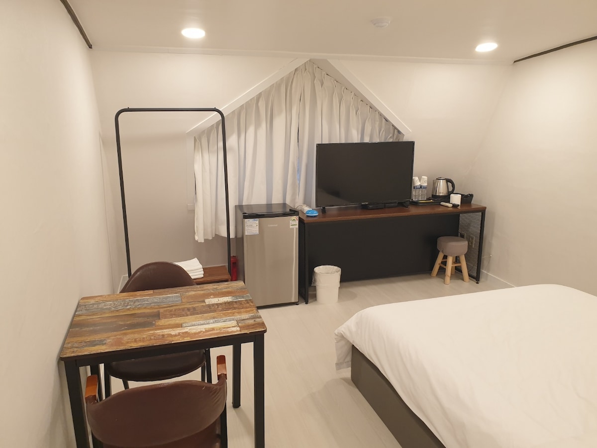 The room features a spacious bed accompanied by a wooden table and two chairs. A large television is mounted on the wall, with water bottles and a kettle on the countertop. A small refrigerator is positioned nearby, adding practical amenities.