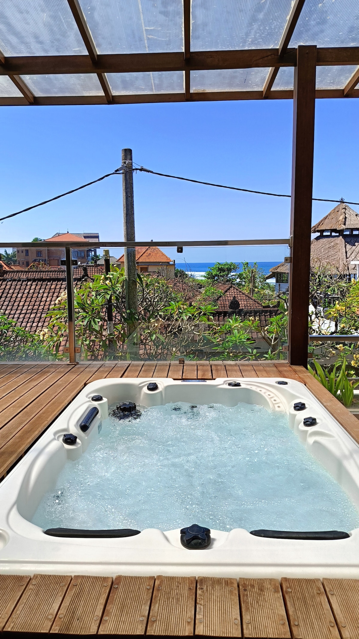 A private jacuzzi is set on a wooden deck, surrounded by greenery and offering a view of the ocean in the distance. The clear water bubbles gently, inviting relaxation, while the clarity of the image captures the bright blue sky above.
