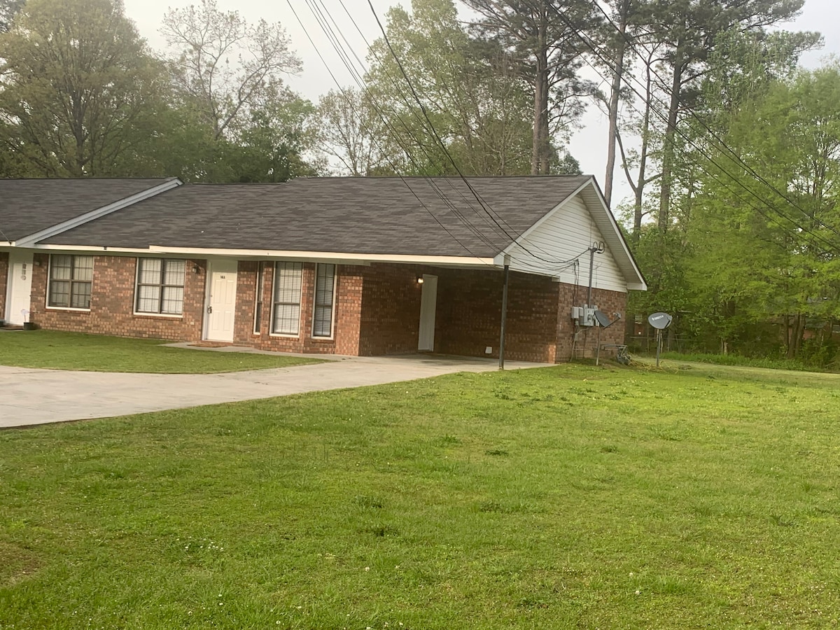 A brick duplex is showcased, featuring a sloped roof and multiple front doors. A spacious driveway is visible, surrounded by a well-kept lawn with green grass and a few trees in the background. Power lines are seen against a light sky.