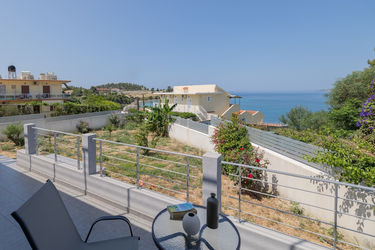 A peaceful outdoor area features a table and chair set, overlooking a grassy yard with various plants. The view includes distant homes and the sea, under a clear blue sky. A relaxed setting invites guests to enjoy the natural surroundings.