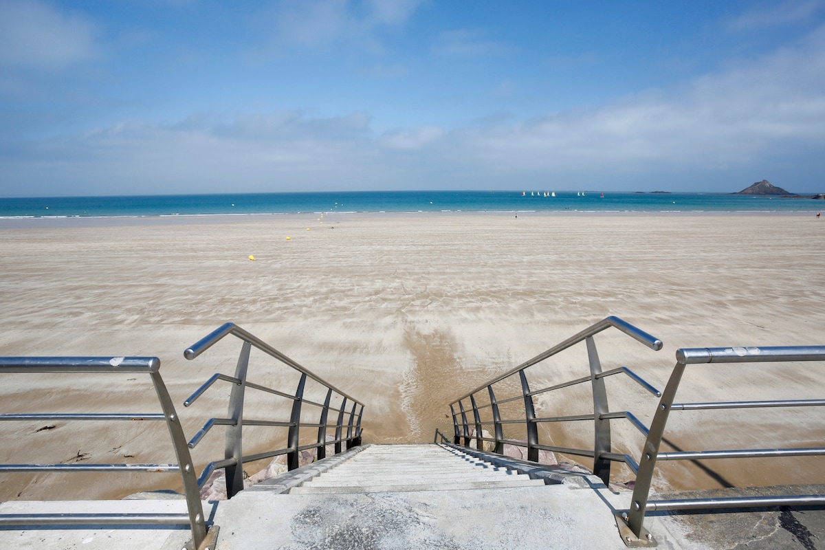 A set of metal stairs leads down to a sandy beach, with the ocean visible in the distance. The shoreline is dotted with colorful buoys, and sailboats can be seen on the calm water under a blue sky with scattered clouds.