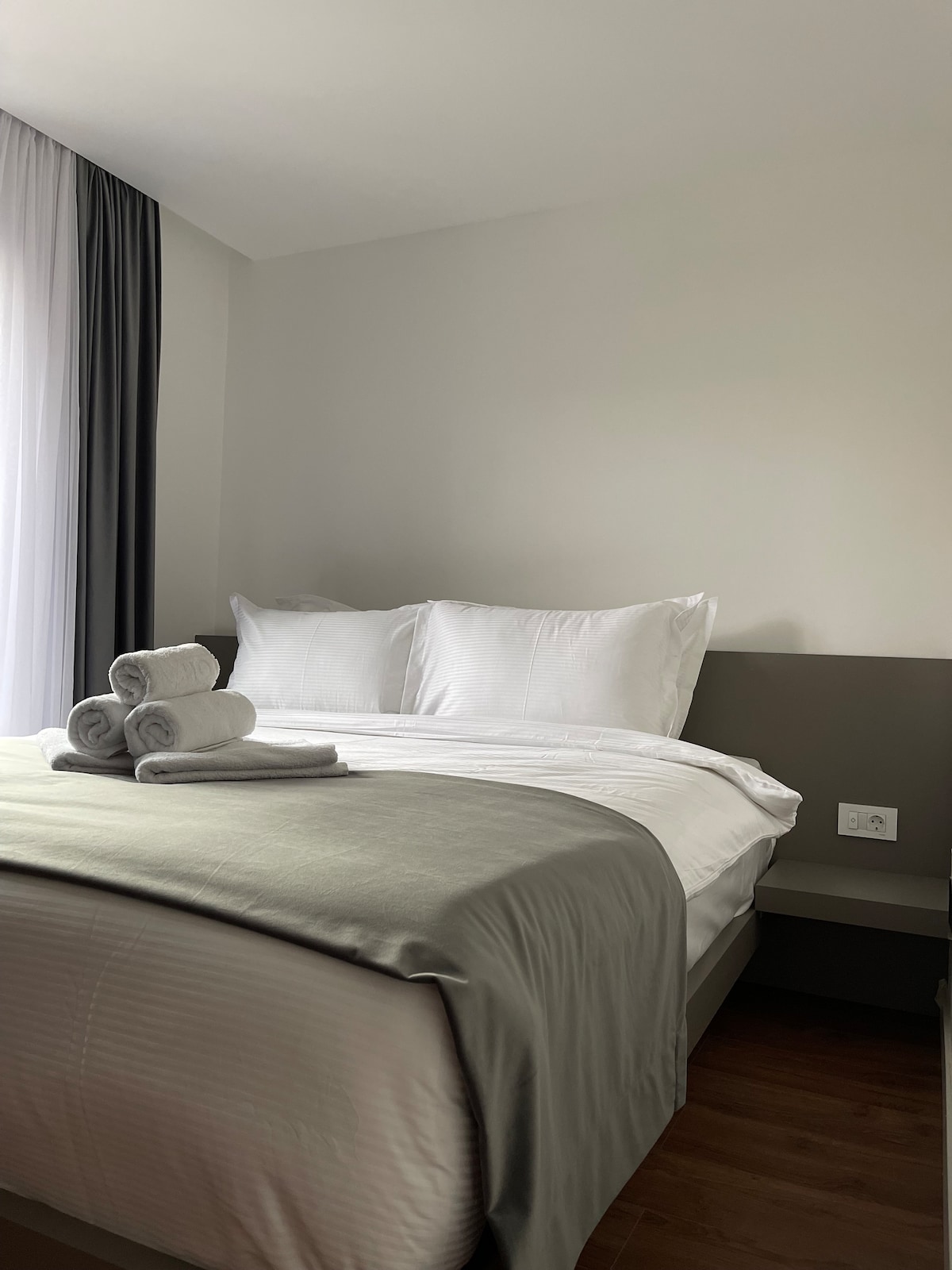 A minimalist bedroom features a spacious bed dressed in white linens and a soft gray blanket. Three neatly rolled towels are arranged on the bed. Natural light filters through sheer curtains, illuminating the neutral color palette.
