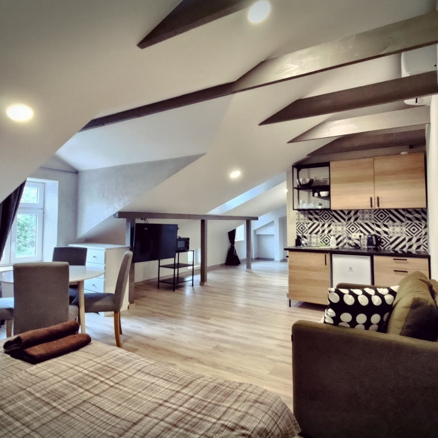 The open-plan living area features a cozy sleeping space, complemented by a stylish kitchenette equipped with essential appliances. A dining table with four chairs is positioned next to the sunny window, while a comfortable seating area is arranged with soft furnishings.