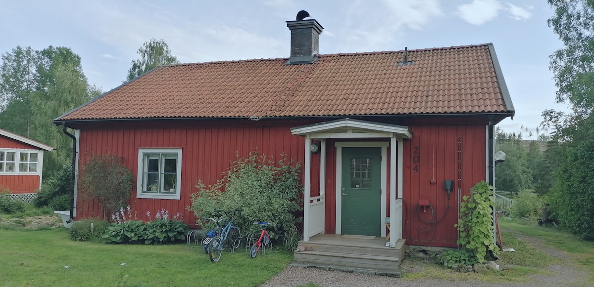 A charming red log house is framed by lush greenery. The entrance features a welcoming porch with a door painted in dark green. A sloped roof with red tiles and a chimney adds rustic character. Bicycles are visible near the entrance, suggesting outdoor activities.