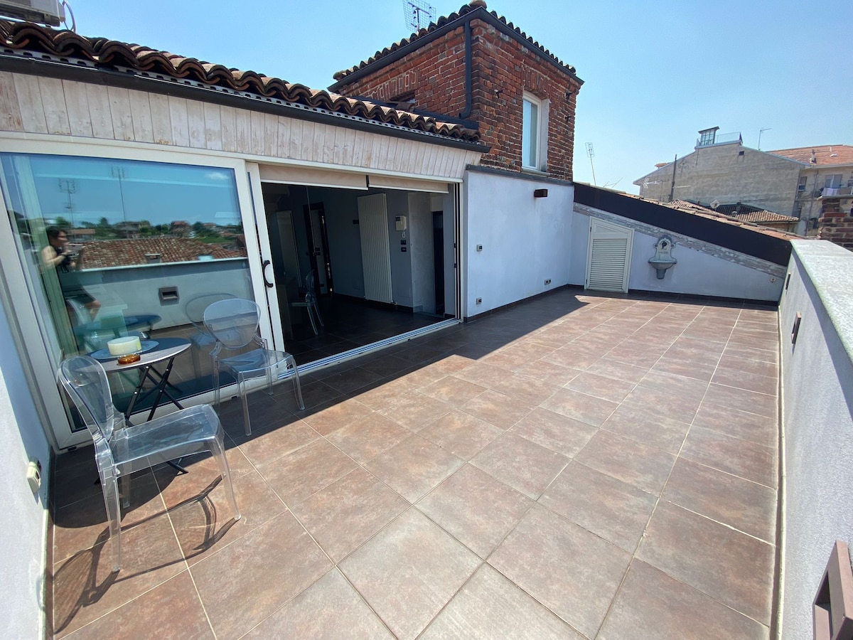 A spacious terrace features modern tiled flooring and clear chairs, offering ample room for outdoor seating. Glass doors leading inside provide natural light. The area is surrounded by a low wall, with a view of the rooftops of San Damiano d'Asti under a clear blue sky.