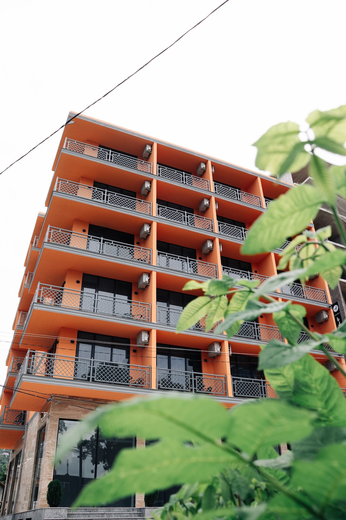 The exterior of a modern multi-story building is shown, featuring large glass windows and metal balconies. The vibrant orange facade contrasts with the greenery in the foreground, providing a contemporary look that frames the structure.