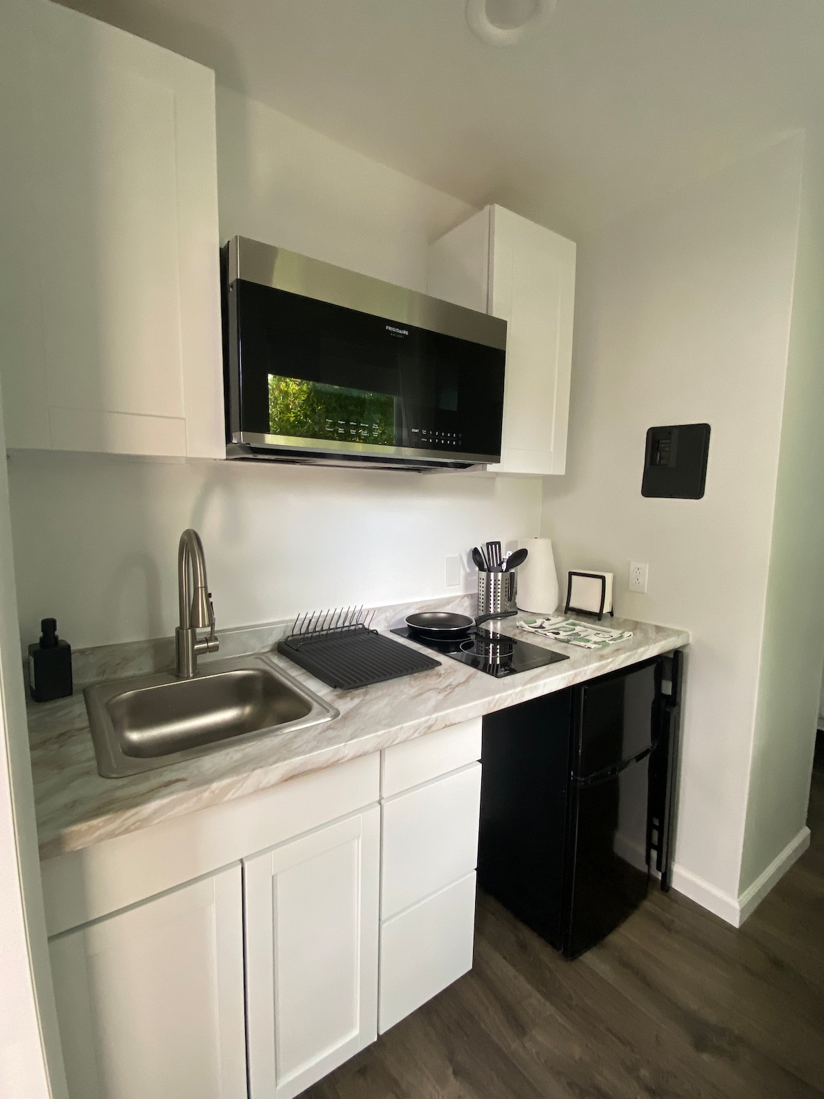 A compact kitchen area is featured, showcasing sleek white cabinetry and a marble countertop. A stainless steel microwave is mounted above a sink, while a small refrigerator is positioned below. Cooking essentials are neatly arranged, creating a functional space for meal preparation.
