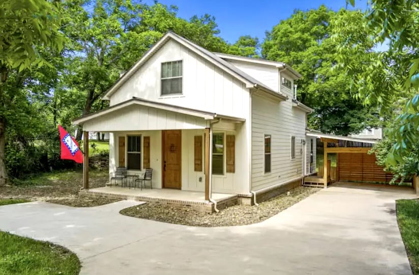 The exterior of a newly-renovated home features a light-colored facade with multiple windows and a front porch. A small seating area with two chairs is visible, alongside a parking space protected by a carport. Lush greenery surrounds the property, creating a serene environment.