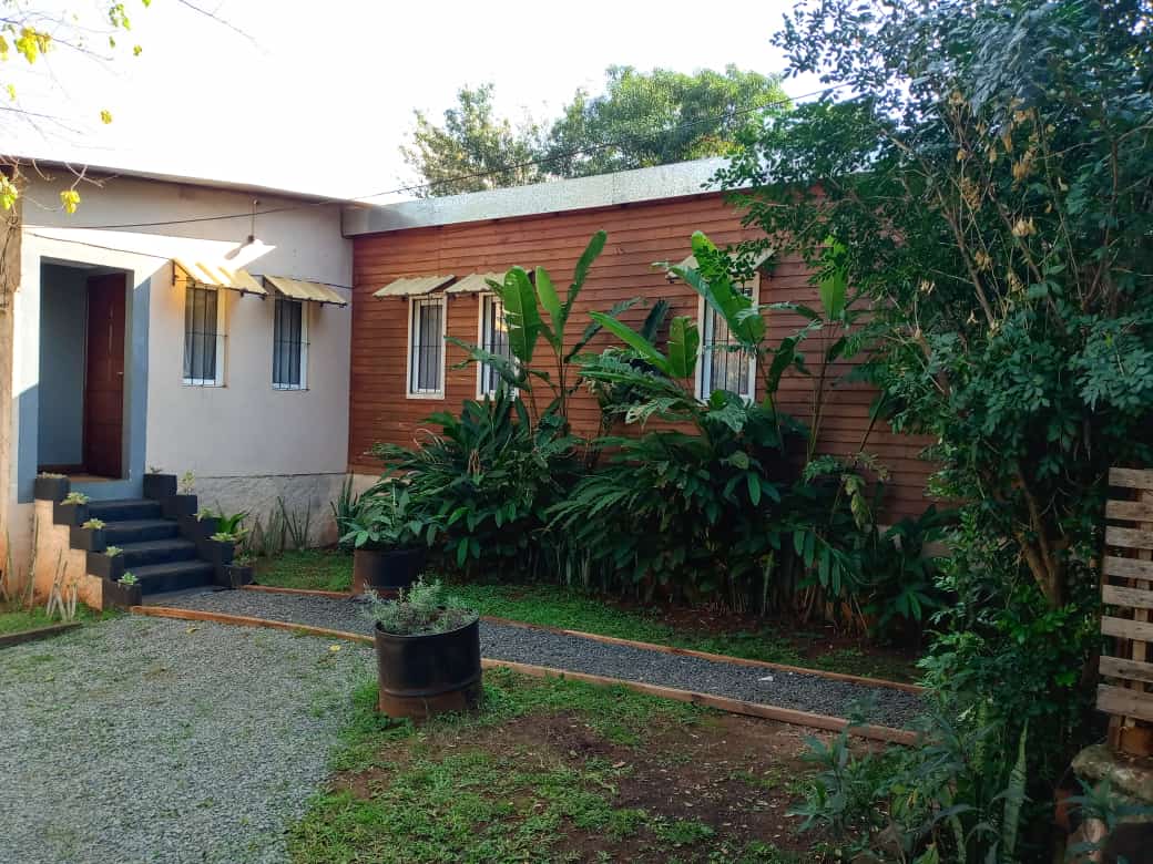 The exterior features a charming wooden house surrounded by lush greenery. Pathways lined with gravel lead to the entrance, while potted plants add natural accents. Sunlight filters through trees, creating a serene outdoor environment.
