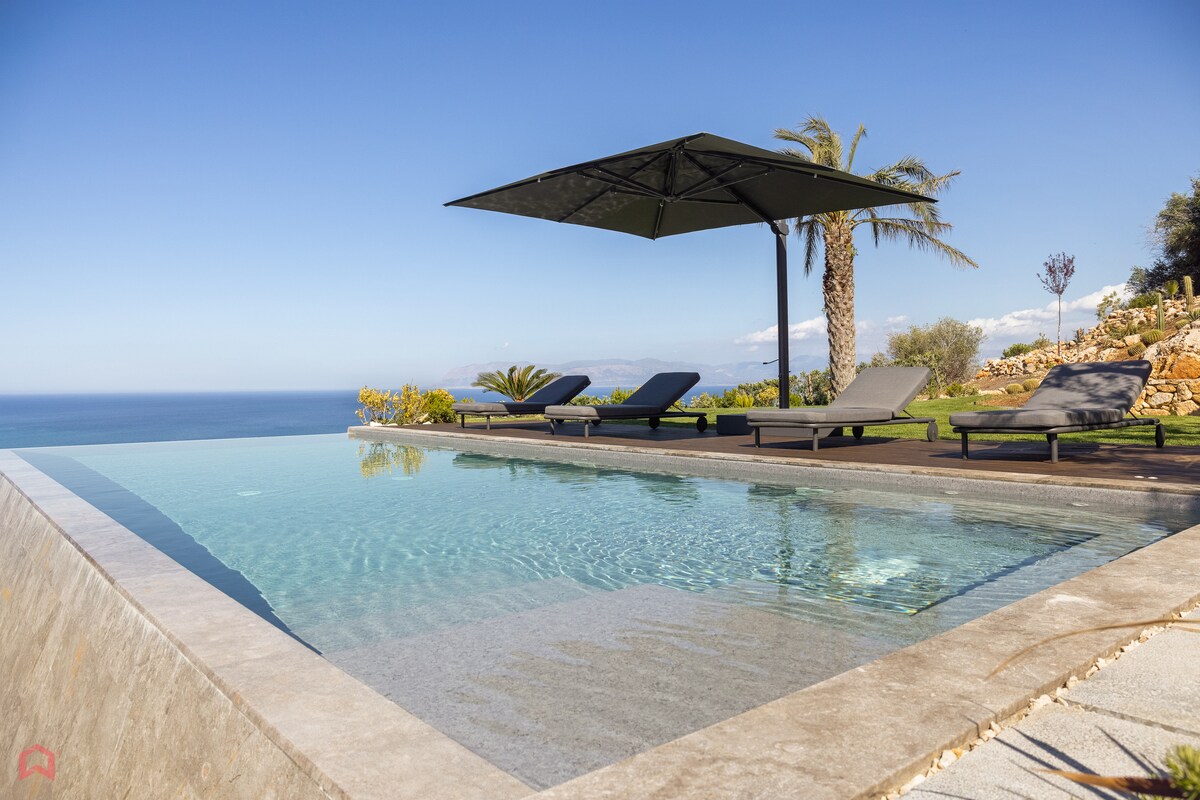 An infinity pool offers expansive views of the coastline and ocean. Lounge chairs are positioned under a large umbrella, providing shade. Palm trees dot the landscape, enhancing the outdoor relaxation area surrounded by vibrant gardens.