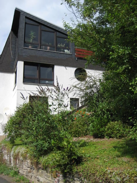 A modern house is surrounded by lush greenery, featuring a combination of dark wood and white exterior. Large windows create an open feel, while a circular window adds unique character. A small garden area with various plants enhances the welcoming atmosphere of the entrance.