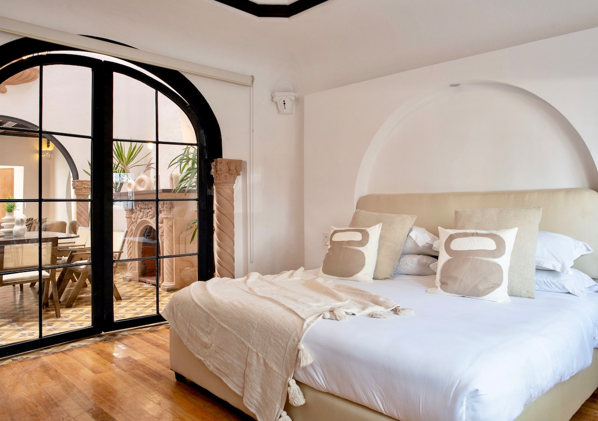 A spacious bedroom is highlighted with a large bed featuring soft linens and decorative pillows. Natural light streams through arched glass doors that lead to a sunny terrace. The neutral color palette and wooden flooring create a serene environment.
