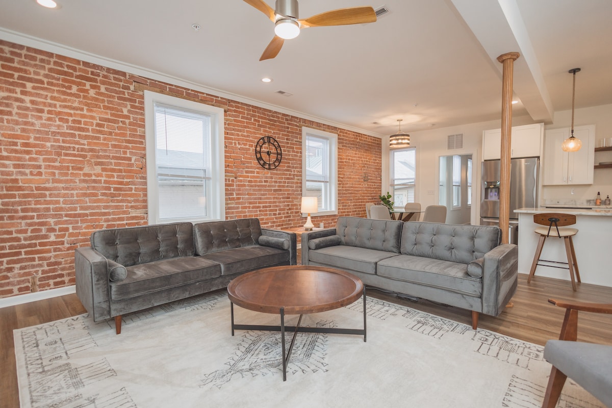 A spacious living area features elegant gray sofas set around a round wooden coffee table on a neutral area rug. Exposed brick walls and large windows provide a warm, inviting ambience, while a dining area and kitchen are visible in the background.