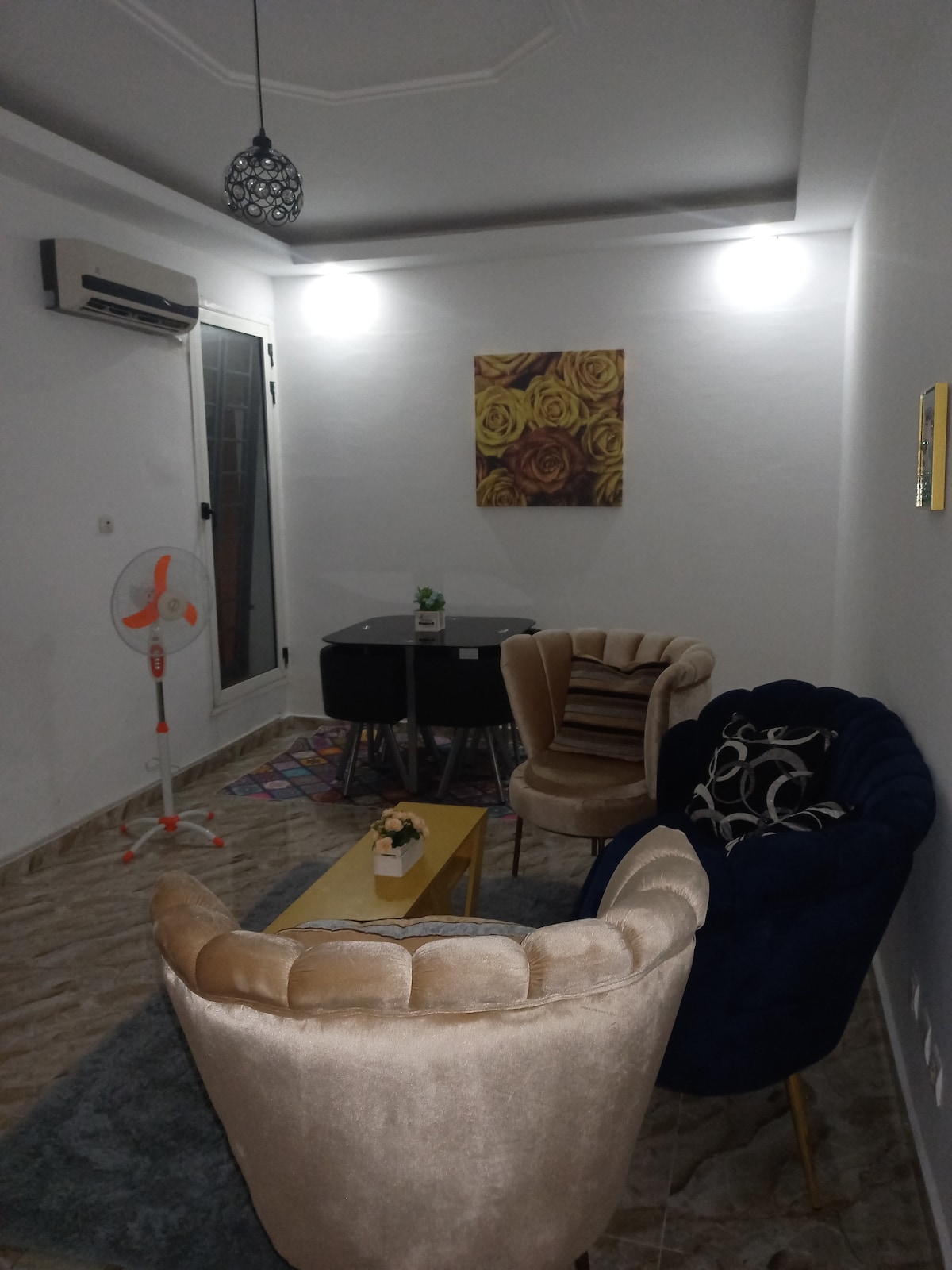 A cozy sitting area features two plush armchairs in contrasting colors and a small table. A glass dining table is placed nearby under warm lighting, and a piece of floral artwork adds a touch of elegance to the wall.