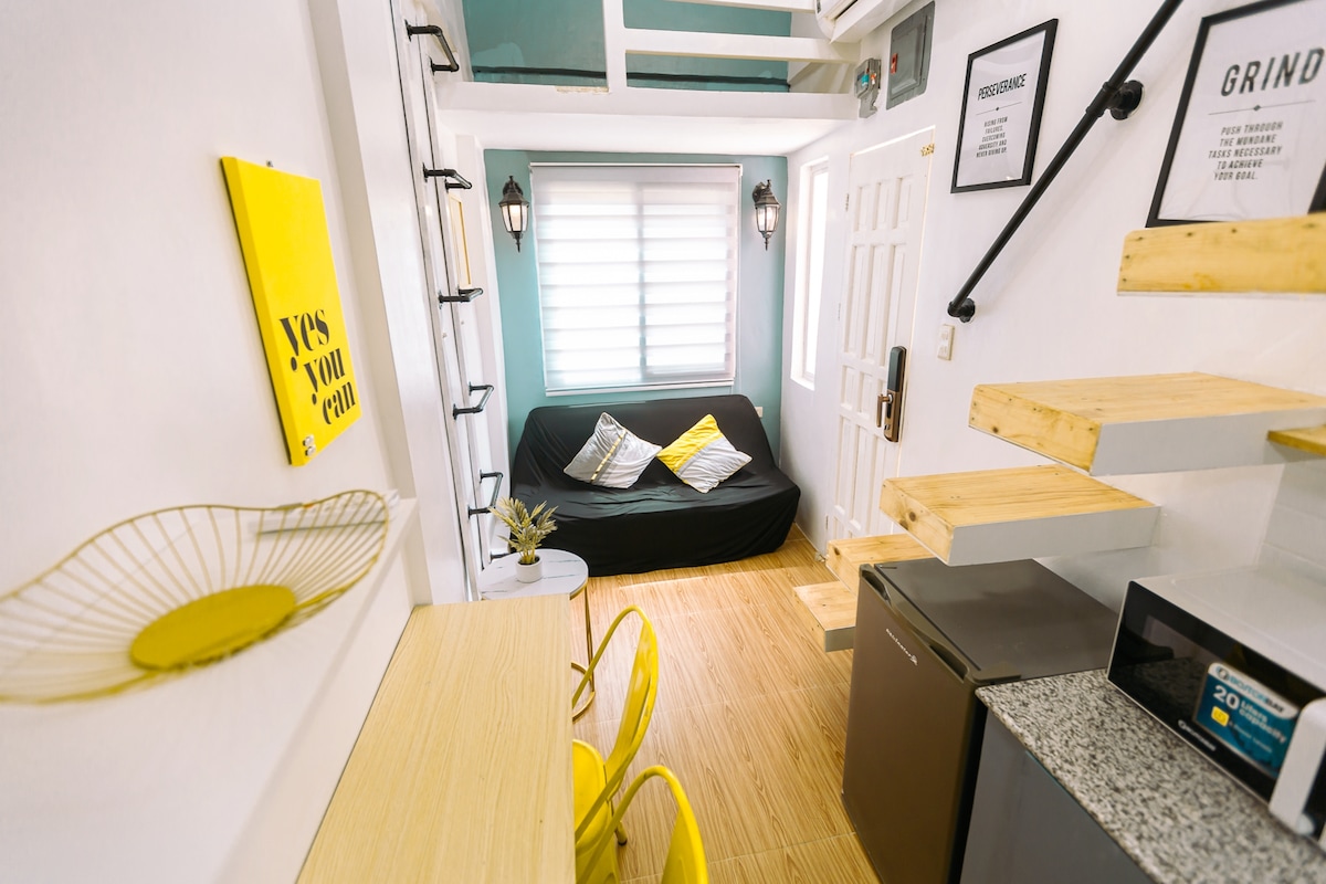 A compact living area features a black sofa adorned with yellow cushions, flanked by windows allowing natural light. A light wood dining table with two yellow chairs is positioned next to a kitchenette, including a mini fridge and microwave, highlighting the efficient use of space.