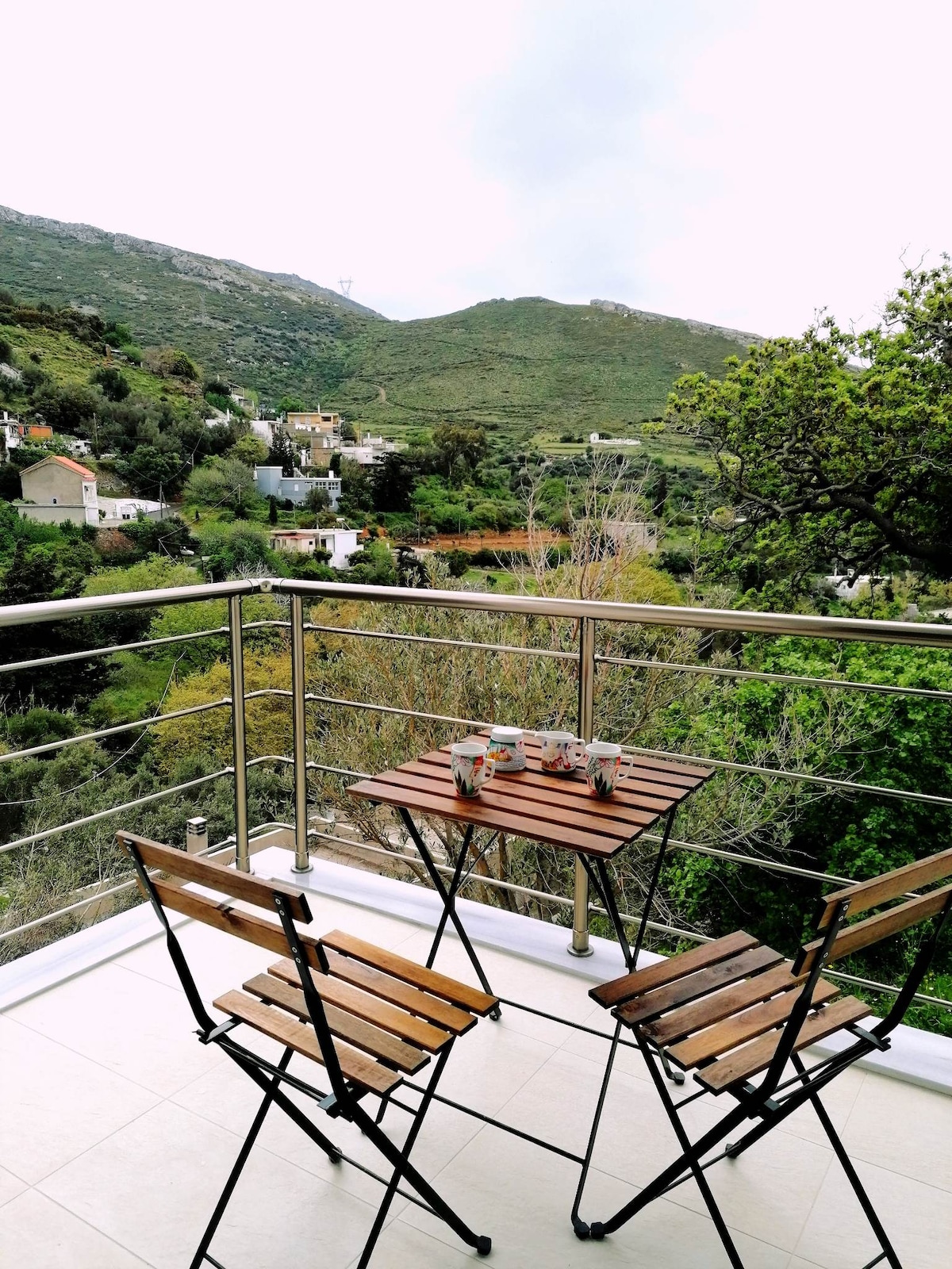 A small balcony features a wooden folding table with two matching chairs, set against a backdrop of lush green hills. The scenic view includes a blend of natural greenery and distant buildings, creating a peaceful atmosphere.