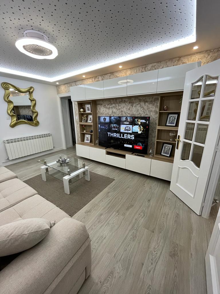 A modern living area features a spacious light-colored sofa and a sleek entertainment unit. A flat-screen television displays a panel of film titles. Soft lighting is provided by a circular ceiling fixture, while decorative elements include a mirrored wall art piece and a glass coffee table.