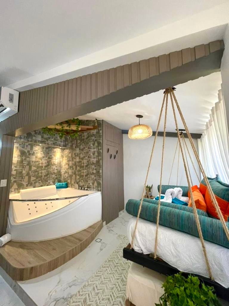 A modern room features a spacious bed adorned with colorful pillows and a textured blanket. A unique suspended seating area complements the design, and a curved, glossy bathtub is positioned nearby. Natural light enters through a large window, enhancing the overall brightness of the space.