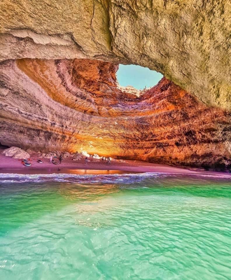 A natural cave is framed by rugged stone walls, revealing a golden sandy beach at its base. Light filters through an opening above, illuminating the vibrant water below. Small figures can be seen enjoying the shoreline, surrounded by tranquil turquoise waves.