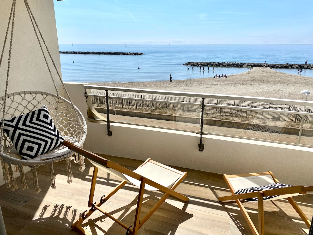 A terrace offers direct views of the calm beach and sea, with two wooden lounge chairs positioned for relaxation. A suspended chair with a geometric cushion adds to the space's comfort and visual interest, while the serene blue water stretches towards the horizon.