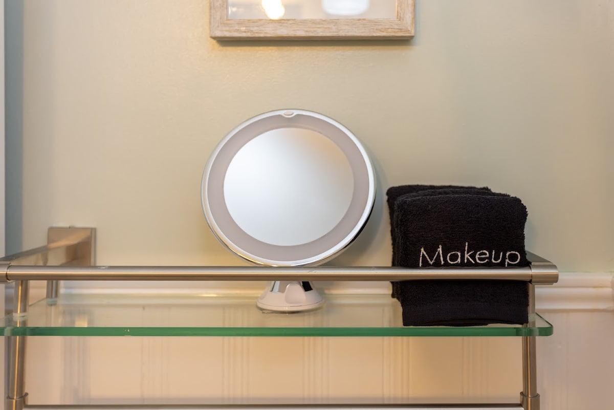 A modern bathroom shelf features a round vanity mirror and a neatly folded black towel labeled 'Makeup'. The shelf is made of glass, supported by a sleek metal frame, contributing to a tidy and organized appearance.