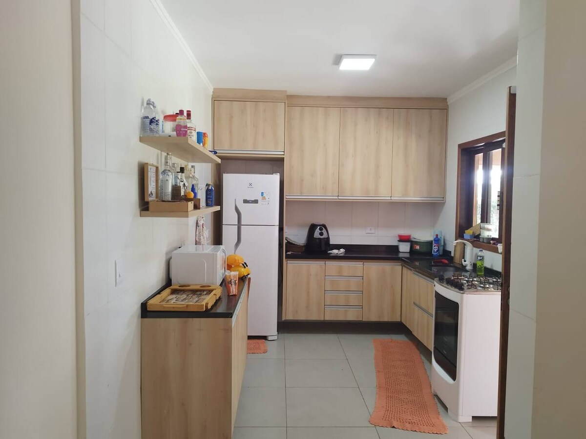 A modern kitchen with wooden cabinetry and a black countertop is showcased. Essential appliances include a refrigerator and a gas stove. Shelves hold various cooking items, and a warm-colored rug adds comfort to the tile floor. Natural light filters through a window.