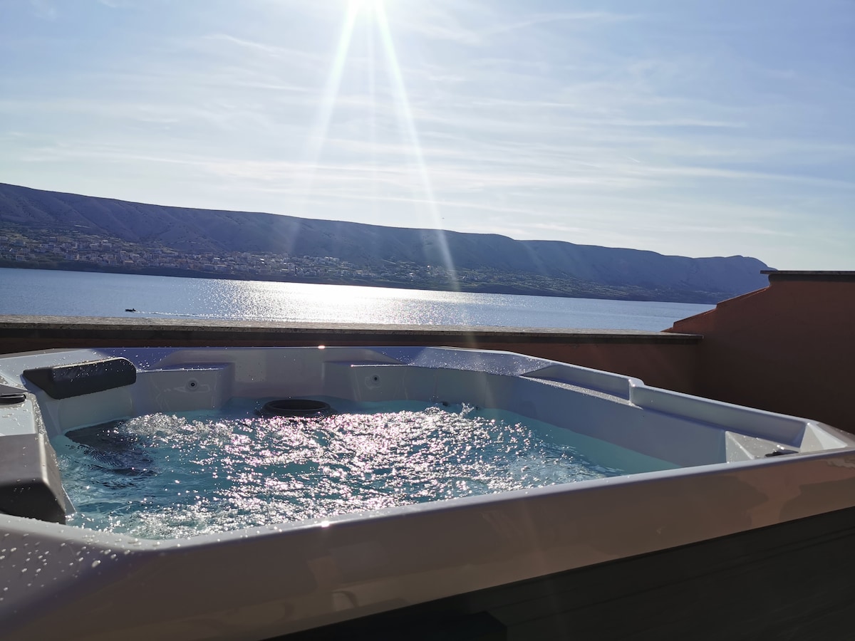 A hot tub filled with clear water is set on a terrace overlooking a scenic lake. Sunlight reflects off the surface of the water, creating a sparkling effect. A backdrop of rolling hills is visible in the distance.