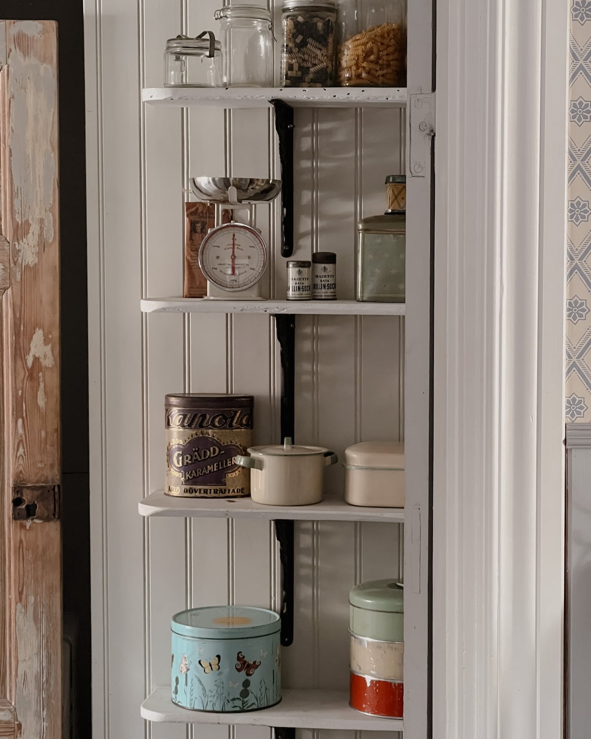 A white shelving unit holds an array of kitchen items, including glass jars filled with ingredients, a vintage scale, and various storage containers. The mix of colors and designs adds character to the space, surrounded by pale wall paneling and patterned tiles.