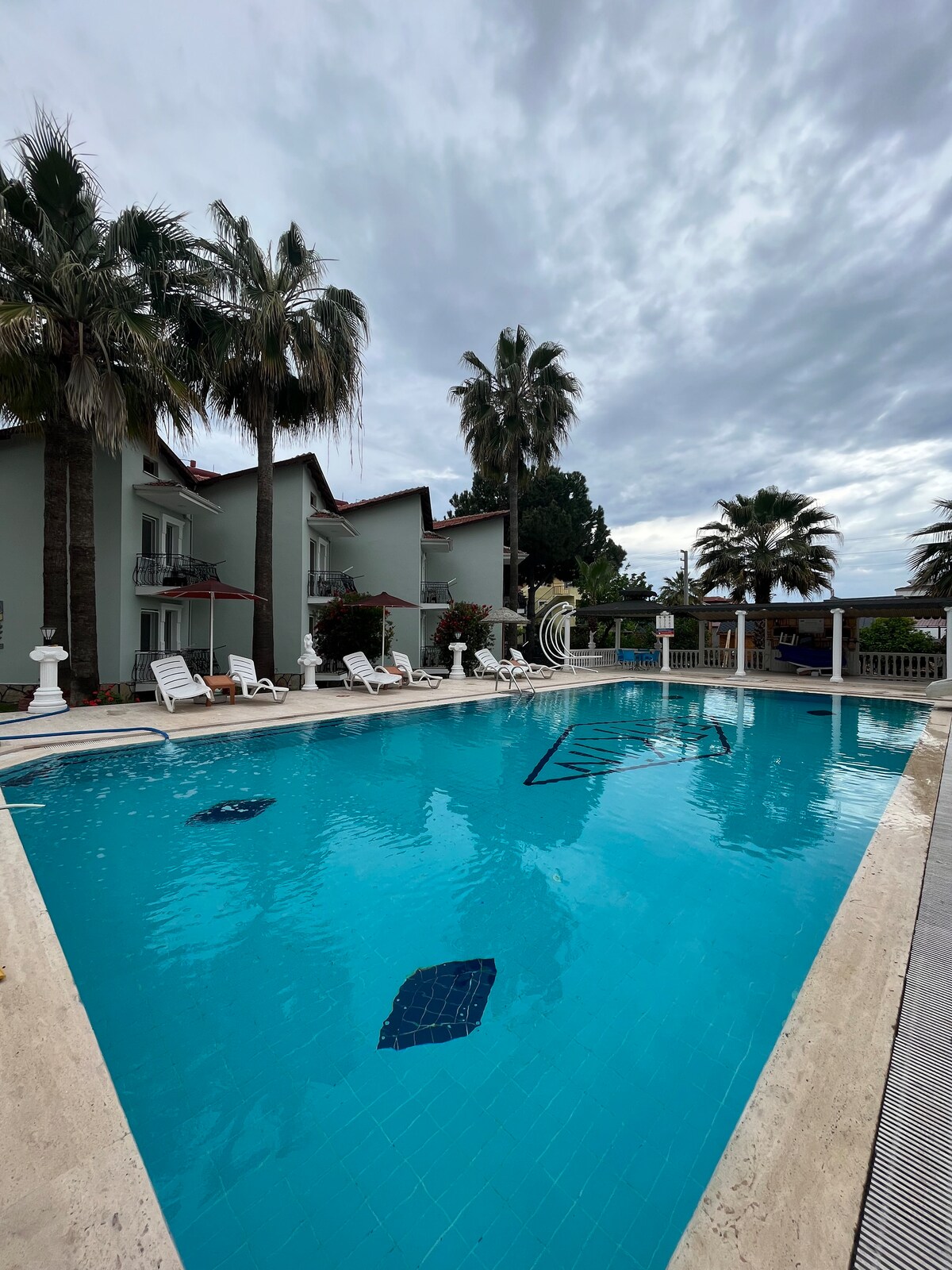 A crystal-clear pool is framed by palm trees and lounge chairs. The surrounding area features a shared space with shaded seating, inviting relaxation. The serene atmosphere is enhanced by the cloudy sky, creating a tranquil environment for guests.