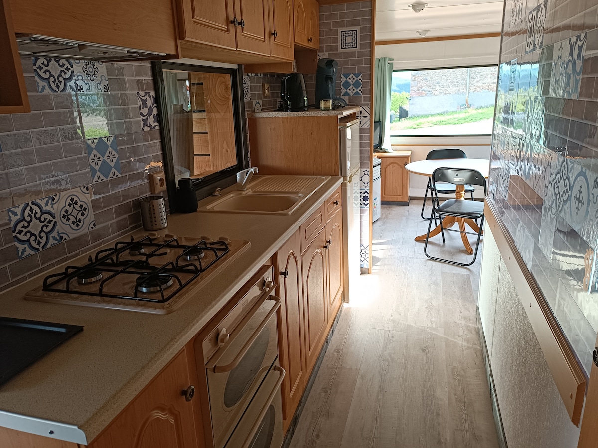 A well-equipped kitchenette is visible, featuring a gas stove, oven, and sink. Light wood cabinetry complements colorful tiled accents. A small dining table with two chairs is positioned near a window, allowing natural light to fill the space.