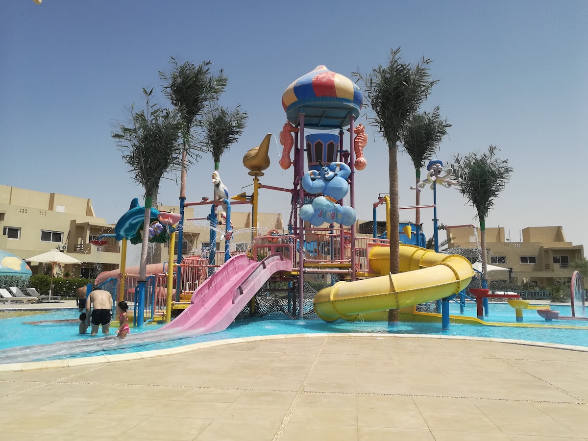 A vibrant water park area features colorful slides and water play structures surrounded by palm trees. The scene captures several visitors enjoying the water in a spacious pool area under a clear blue sky.