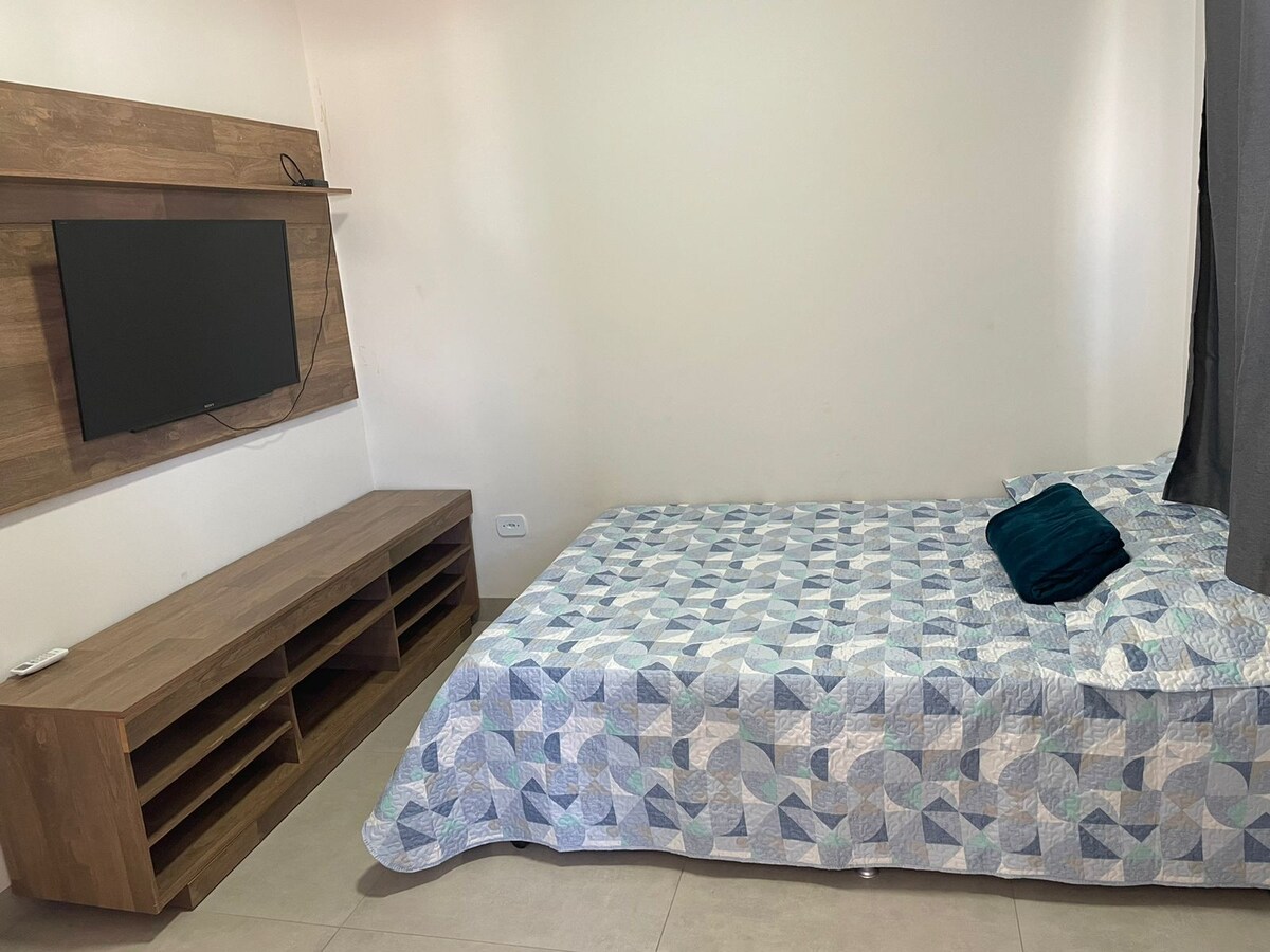 The sleeping area features a spacious bed covered with a patterned quilt. A teal pillow adds a pop of color. A flat-screen television is mounted on the wall above a wooden media console with shelving, enhancing the simplicity of the space.