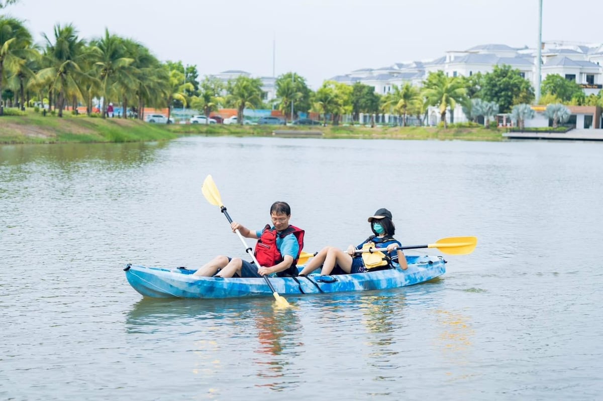 A kayak is seen gliding smoothly across a calm water surface. Two individuals are seated in the kayak, each holding a paddle. Lush greenery lines the bank of the water, and modern residential buildings can be observed in the background.