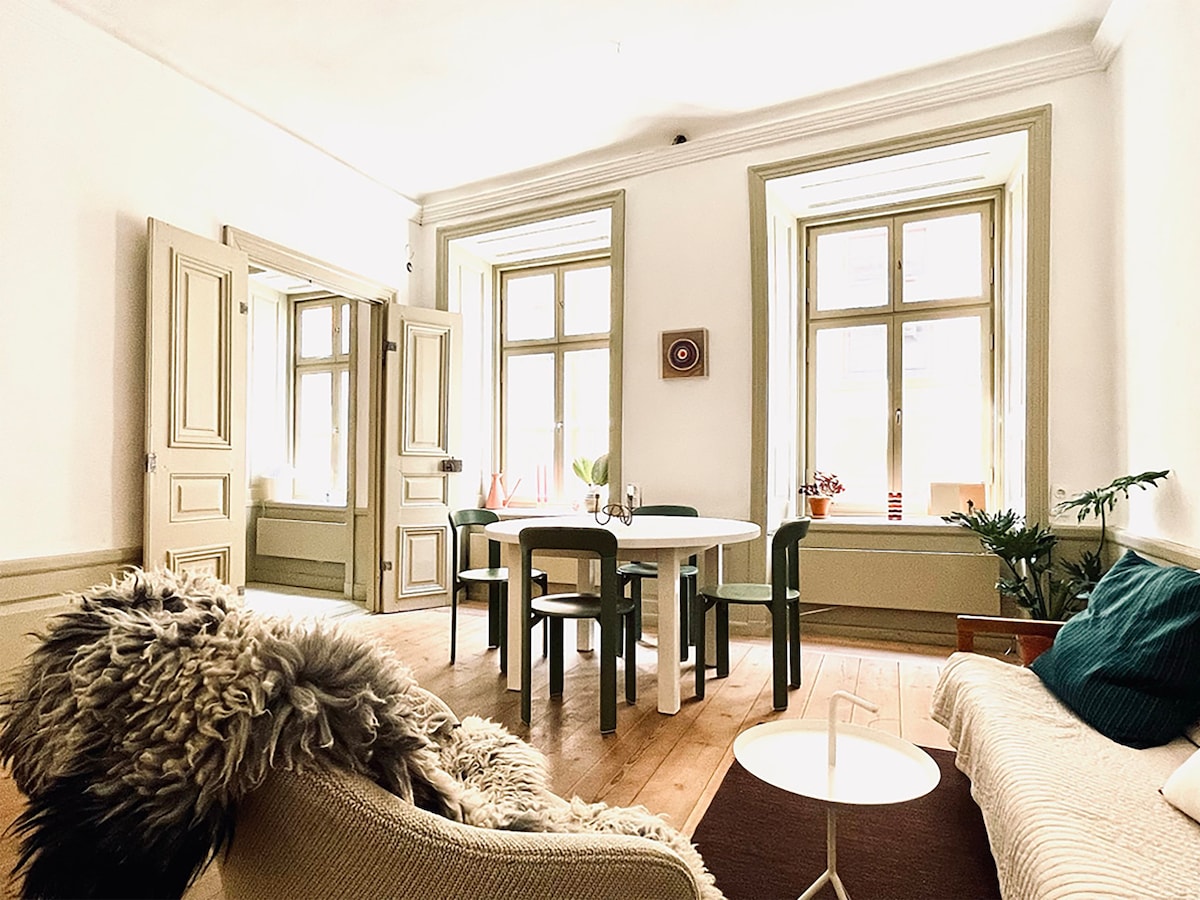 A spacious living/dining room features large windows allowing natural light to fill the area. A round dining table is accompanied by green chairs, while a cozy sofa and soft textile accents provide comfort. Original wall details highlight the historic charm of the apartment.