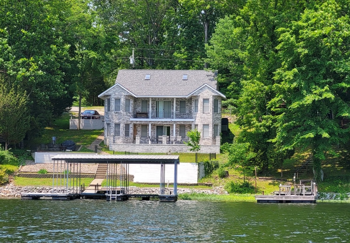 A charming lakefront home is situated among lush greenery, showcasing a spacious two-story structure with a wide porch. A double dock can be seen at the water's edge, providing access for boating and fishing activities. The landscaped yard features steps leading to the shore.