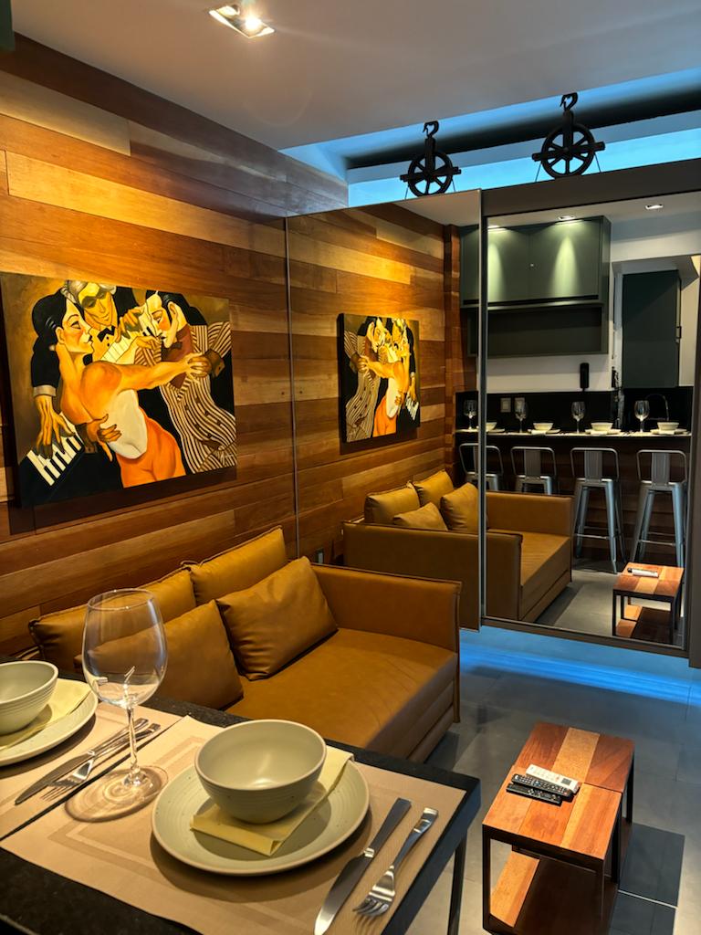 A cozy living space features wooden paneling and vibrant artwork. A brown sectional sofa is positioned beside a dining setup with plates and glasses. The kitchen area is visible in the background, fitted with barstools along a countertop.